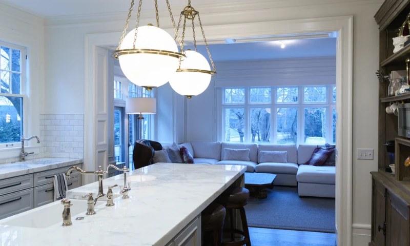 A modern kitchen in East Grand Rapids, MI, with marble countertops and brass light fixtures, offers a stylish refresh, opening to a living area with a large sectional sofa, coffee table, and windows overlooking trees.