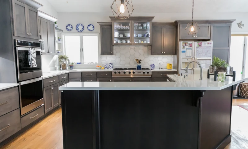 A modern kitchen in Cascade, Michigan, with dark cabinets, white countertops, stainless steel appliances, and open shelving with dishes. Pendant lights over a central island refresh the space with a stylish touch.