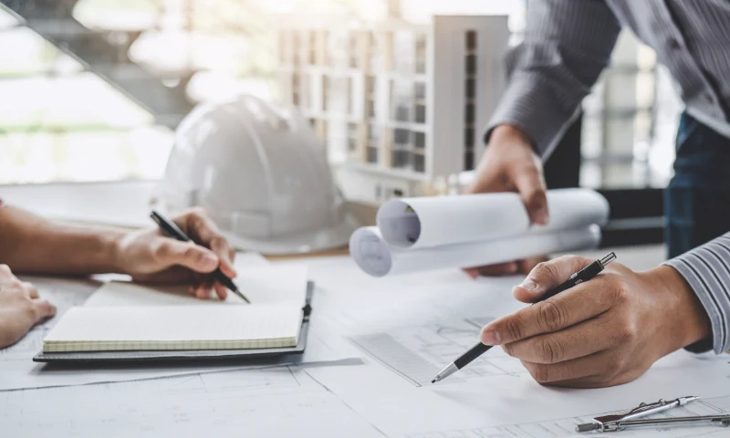 Two people refresh architectural blueprints at a desk with pens and notepads; a white construction helmet and building models are in the background.