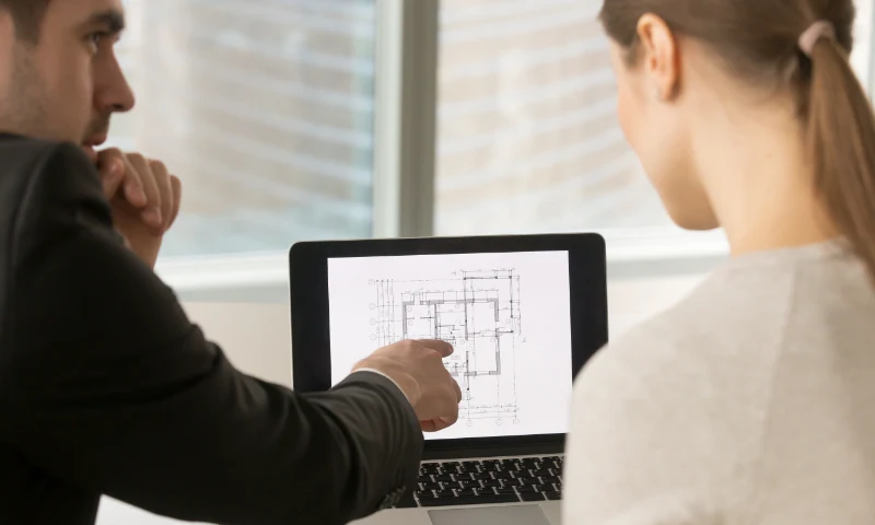 Two people looking at a laptop screen displaying a technical architectural floor plan, with one person pointing at the screen to suggest a refresh of the current design.