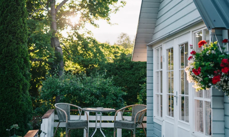A small outdoor patio with two wicker chairs and a round table offers a cozy spot to refresh beside a light blue house with white trim and red flowers by the window, surrounded by trees and greenery.