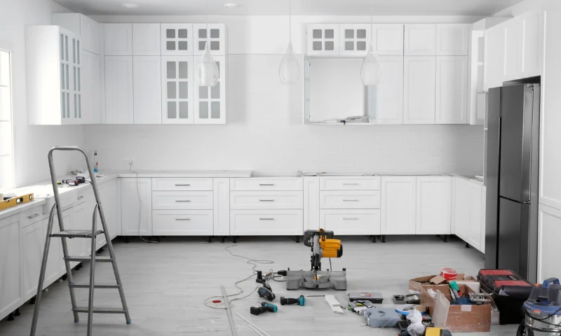 A modern white kitchen in West Michigan gets a refresh, with tools, a ladder, and construction materials scattered on the floor. Cabinets and appliances are installed but remain unfinished in this renovation scene.