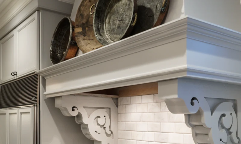 Large metal pans are displayed on a white kitchen shelf above a stove, with decorative wood supports and a white tile backsplash for a classic refresh, in East Grand Rapids, Michigan.