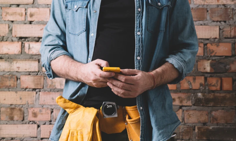 Person wearing a denim shirt and a tool belt stands in front of a brick wall, using a yellow smartphone to refresh project plans.