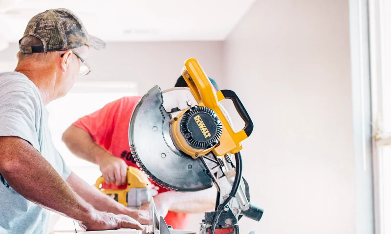 Two people use power tools and a circular saw to cut wood indoors, refreshing their space as sunlight streams through a window in the background.