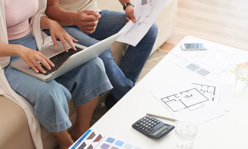 Two people sit on a couch looking at a laptop and house plans, surrounded by papers, a calculator, and color swatches on a white table as they plan to refresh their living space.