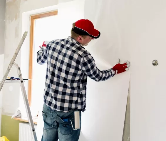 A person in a plaid shirt and red cap is refreshing a wall with new wallpaper, using a smoothing tool while standing next to a ladder.