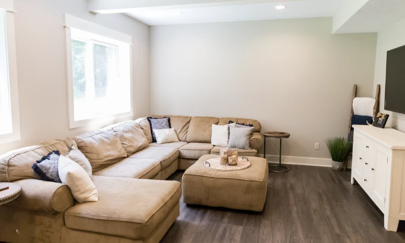 A living room refresh in Sparta, MI, with a beige sectional sofa, matching ottoman, small round table, white cabinet, decorative items, and wood-look flooring. Light streams in through two windows.