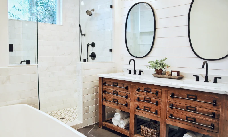 A modern bathroom in Grand Rapids, MI, with a glass shower, dual sinks with black fixtures, two oval mirrors, and a wooden vanity with drawers and rolled towels underneath—perfect for a quick refresh.