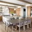 A modern kitchen in West Michigan, with wooden cabinets, a large island, a gray dining table for six, glass pendant lights, open shelving, and large windows that refresh the space with views of lush outdoor greenery.