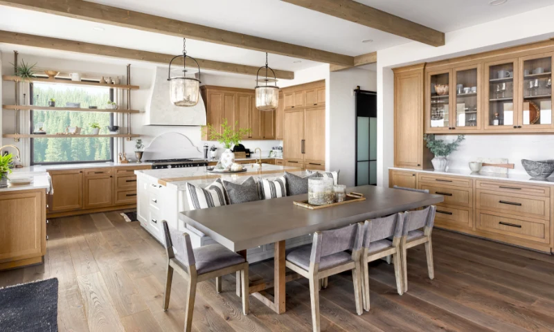 A modern kitchen in West Michigan, with wooden cabinets, a large island, a gray dining table for six, glass pendant lights, open shelving, and large windows that refresh the space with views of lush outdoor greenery.