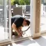 A man kneels on the floor, refreshing a sliding glass door that leads to an outdoor patio area with furniture.