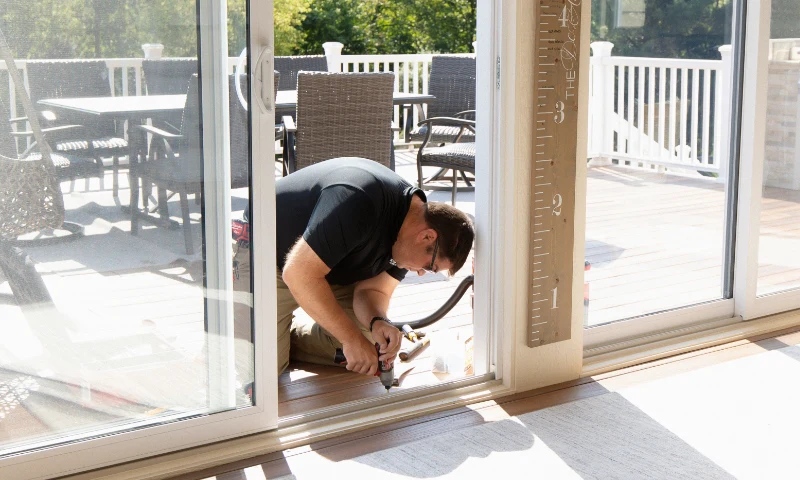 A man kneels on the floor, refreshing a sliding glass door that leads to an outdoor patio area with furniture.