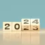 Four wooden blocks display the numbers "2023," with the last block rotated to refresh the year from 2023 to 2024, set against a plain blue background.