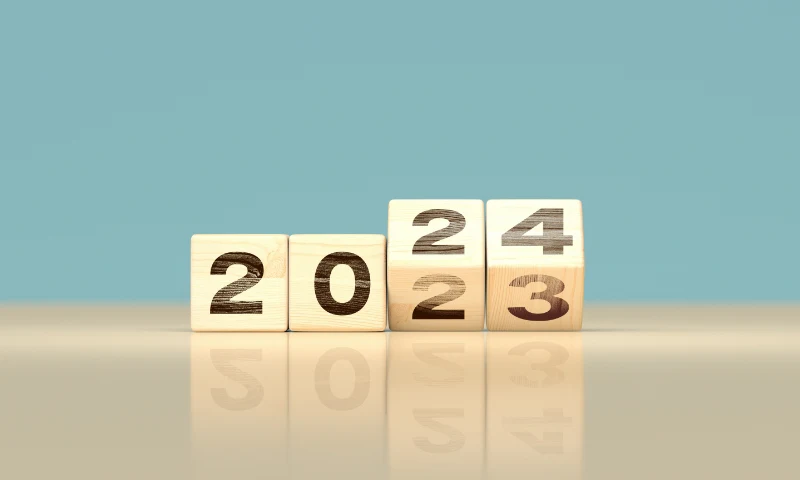 Four wooden blocks display the numbers "2023," with the last block rotated to refresh the year from 2023 to 2024, set against a plain blue background.