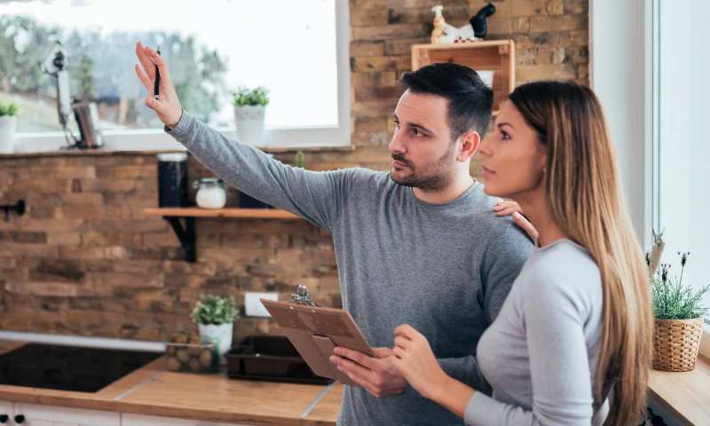 A man holding a clipboard points at something off screen while standing next to a woman in a modern kitchen with brick walls and wooden shelves, discussing ideas to refresh the space.