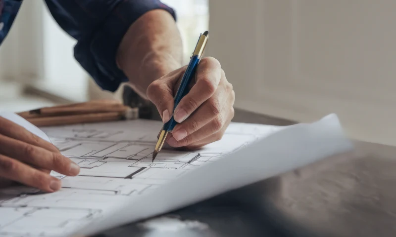 A person’s hand draws lines on a technical blueprint with a pen, giving a refresh to the design, while architectural tools are visible in the background on a desk.