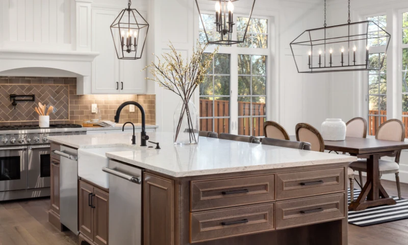 A modern kitchen in West Michigan, with a large island, stainless steel appliances, pendant lighting, and white cabinets, offers a fresh, refreshed feel, complemented by a dining area featuring a wooden table and upholstered chairs.