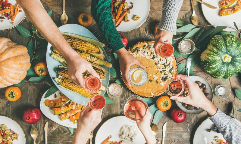 Five people refresh with drinks over a table set with autumn vegetables, corn, pie, and pumpkins, suggesting a festive meal or holiday gathering.