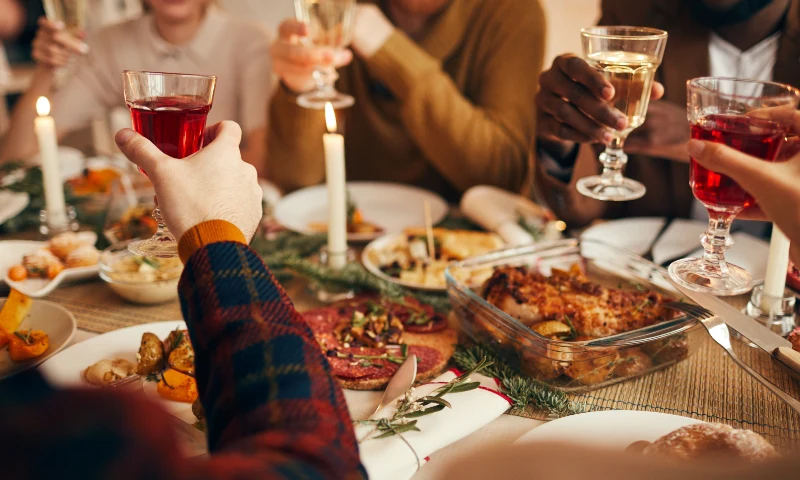 A group of people sit around a dining table, holding up glasses for a toast, enjoying a refresh of good company, with various dishes and candles placed on the table.