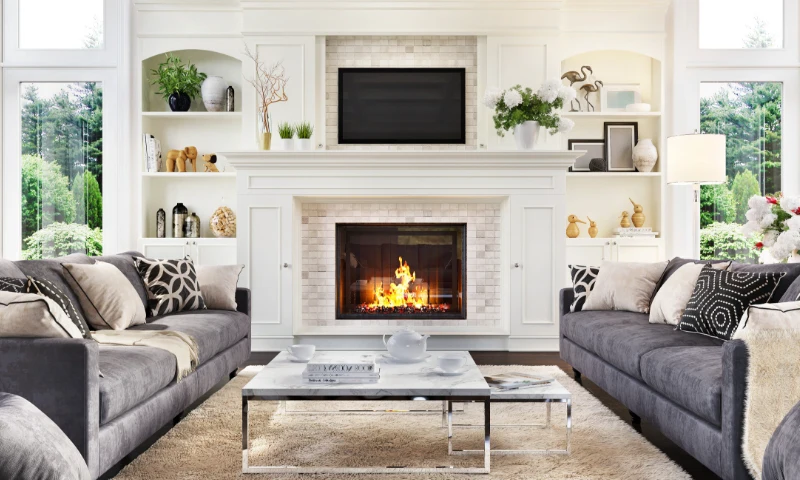 A modern living room in West Michigan, with two gray sofas, a coffee table, and a lit fireplace beneath a wall-mounted TV, surrounded by built-in shelves with decor and large windows that refresh the space with views of greenery outside.