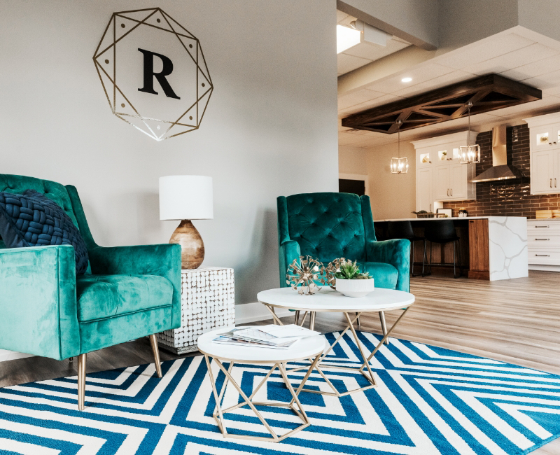 Modern living room with two teal armchairs, a white lamp on a side table, round white coffee tables, and a geometric blue rug at the Ridgewood Home Construction showroom in Grand Rapids, Michigan.