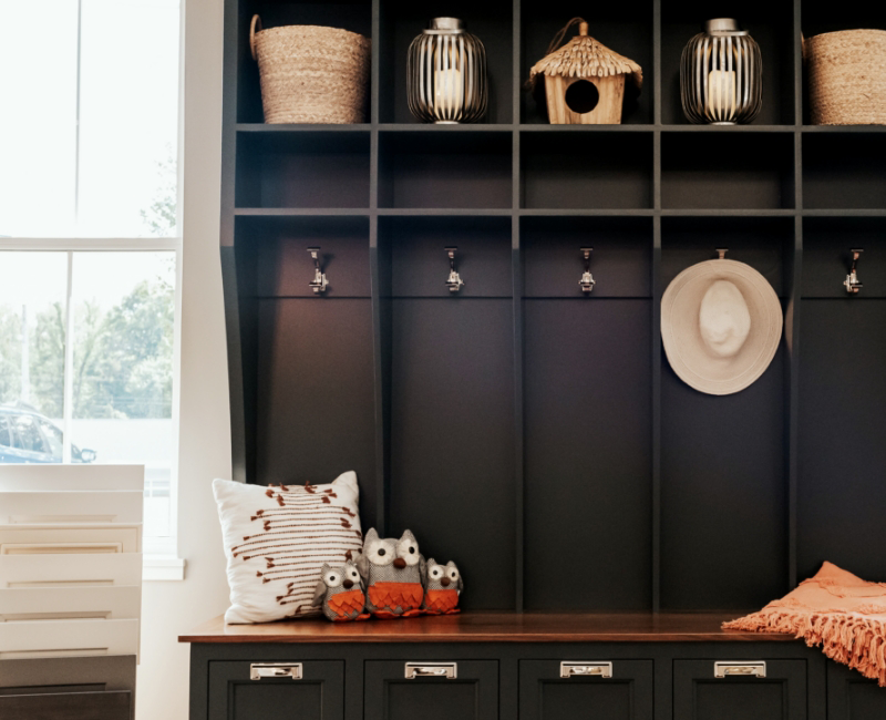 A dark built-in cubby organizer in Grand Rapids, MI, with hooks, woven baskets, a birdhouse, lanterns, a striped pillow, plush owl toys, and a hat hanging on a hook sits by the window—perfect for showcasing your remodeling process.