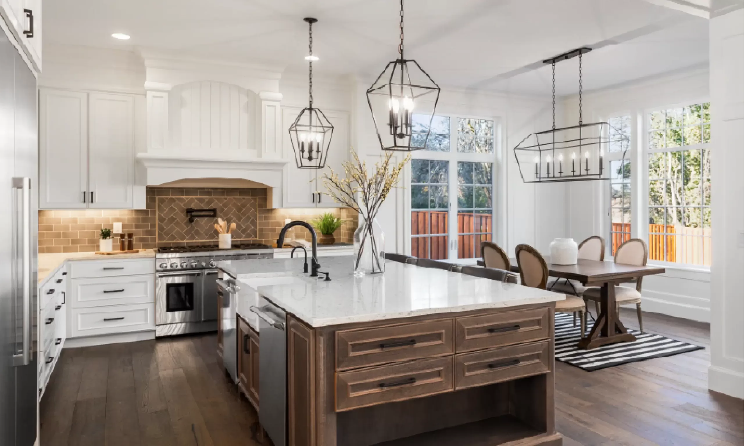 A modern kitchen in West Michigan, with white cabinets, a large island with a sink, stainless steel appliances, and pendant lights showcases the latest kitchen design trends. An adjacent dining area features a wooden table and upholstered chairs.