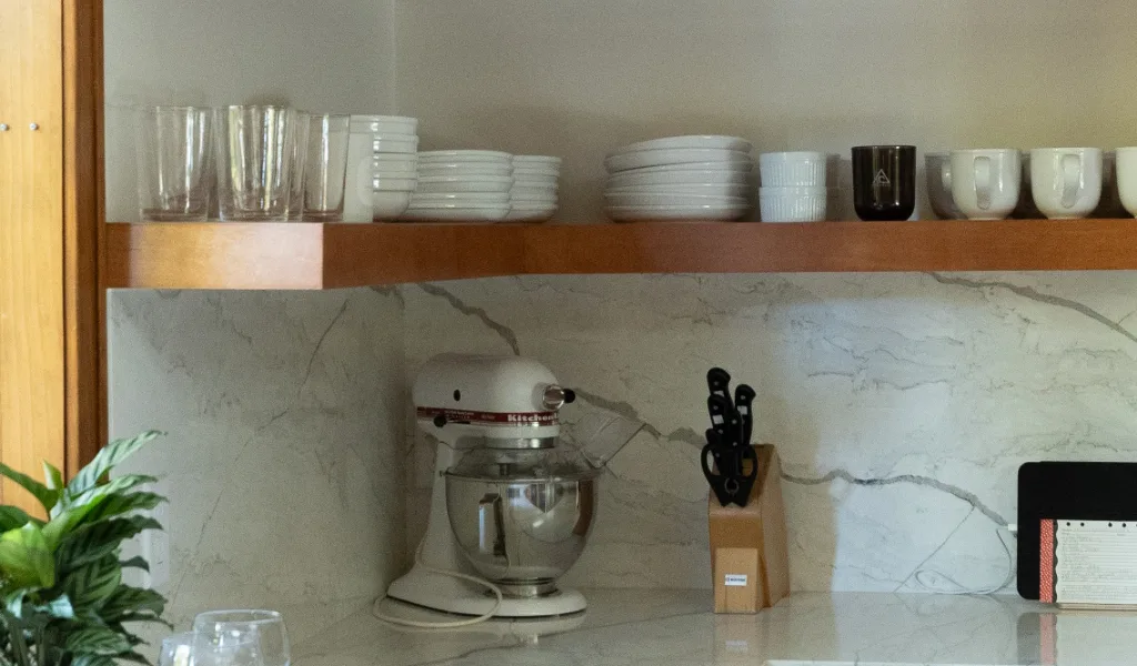 A kitchen counter from a Rockford kitchen remodel, with a stand mixer, knife block, potted plant, tray with glasses and pumpkins, and shelves holding dishes, mugs, glasses, and stacked books.