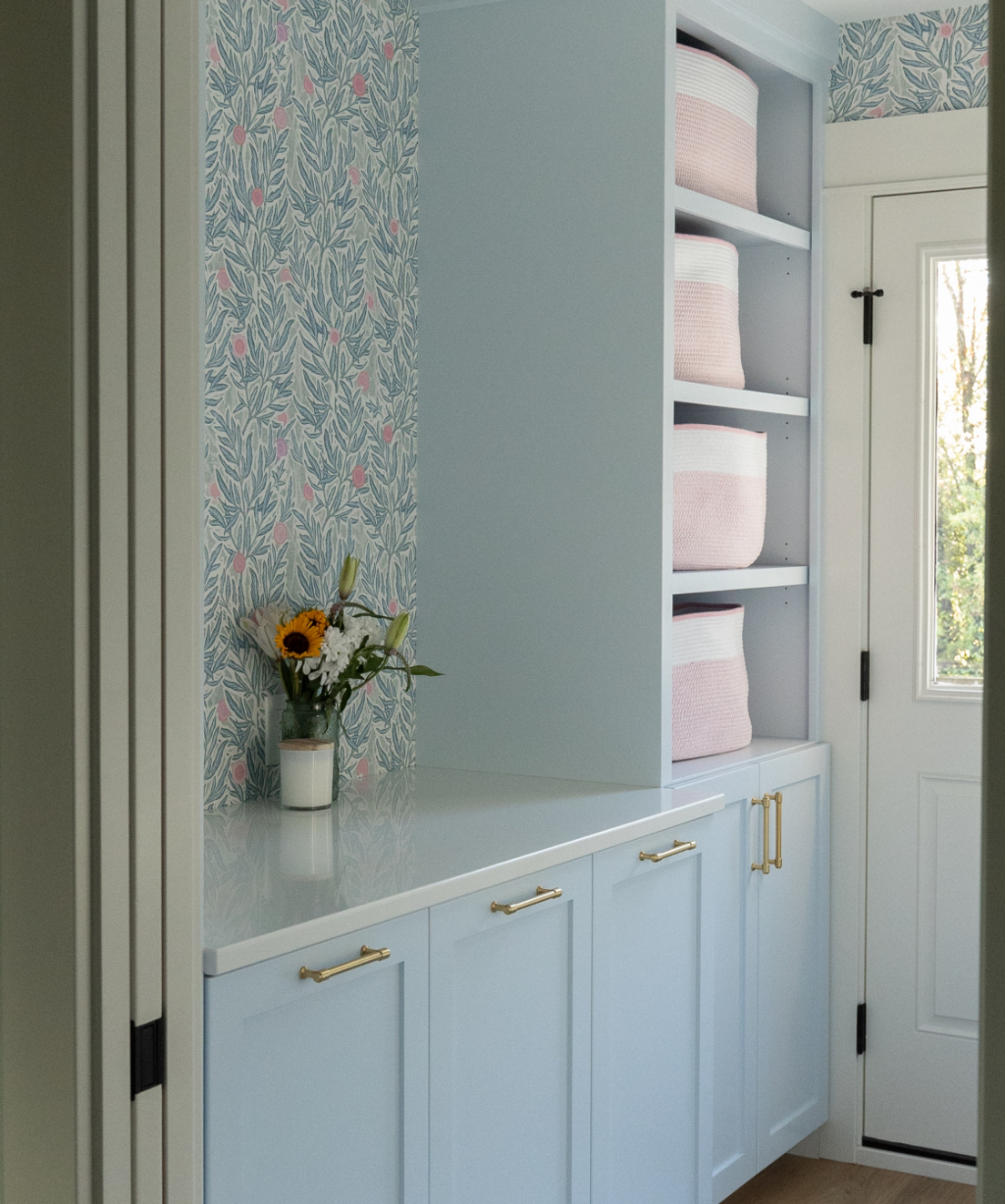 Satkoski – 1 A light blue mudroom from a Byron Center Whole Home Remodel features gold hardware, floral wallpaper, a vase of flowers on the counter, and open shelves holding pink-striped baskets.