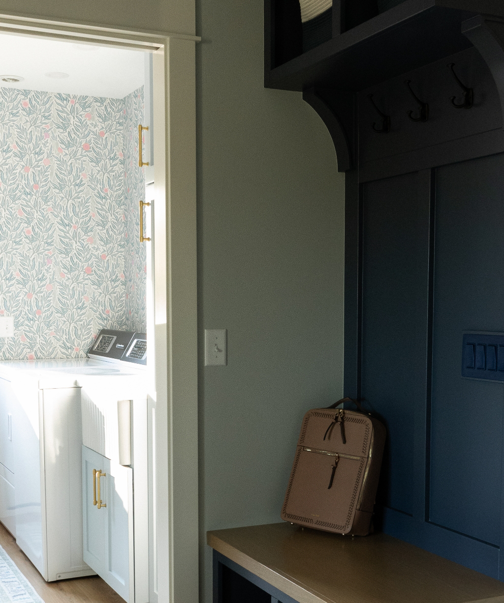 A mudroom with dark blue built-in storage, coat hooks, a beige bench, and a tan bag; through the doorway, the Byron Center Whole Home Remodel reveals a laundry room with floral wallpaper.
