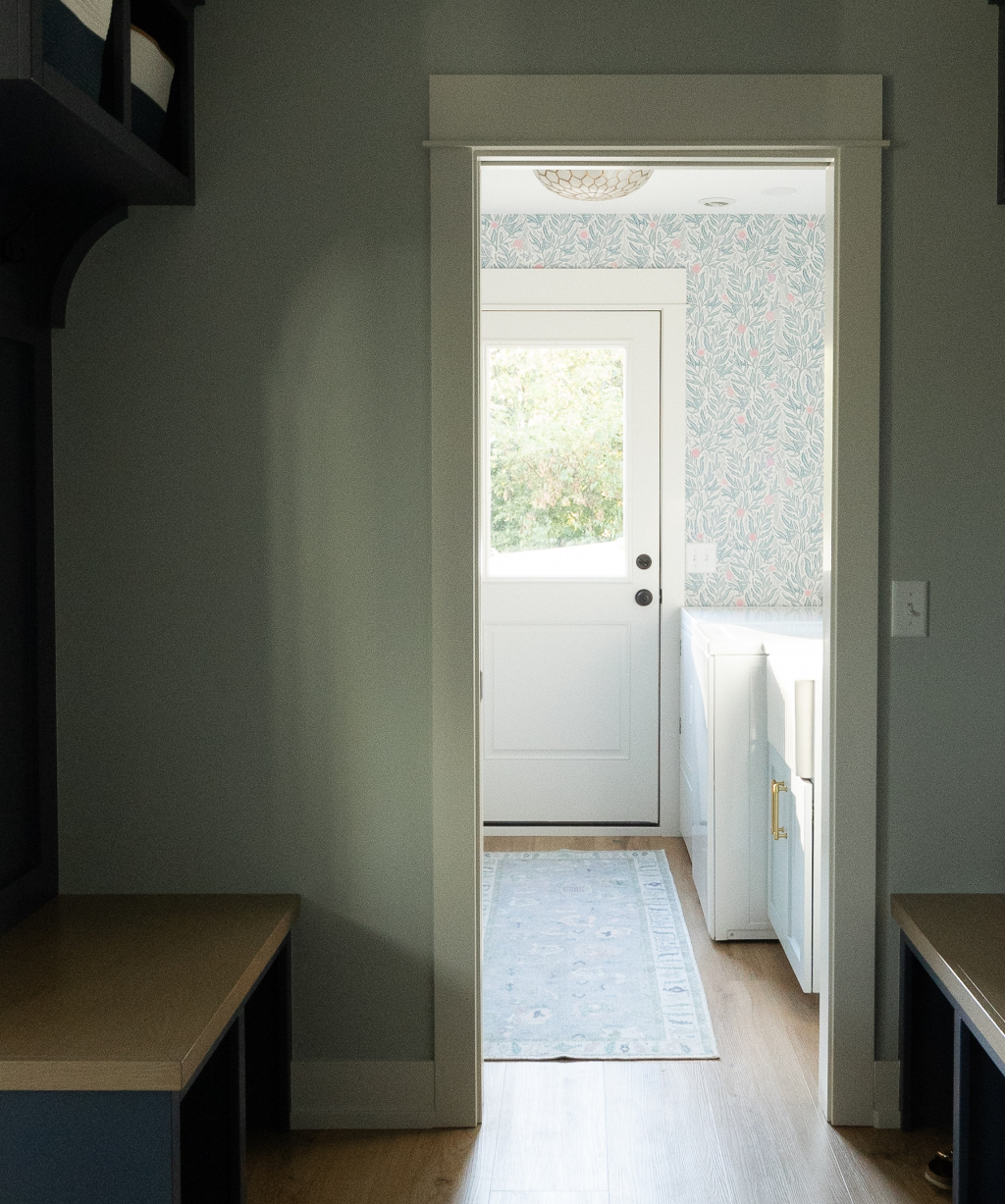 A view of a white door with a window, located at the end of a hallway with built-in benches on either side, highlights this Byron Center Whole Home Remodel and the patterned wallpaper in the adjoining room.