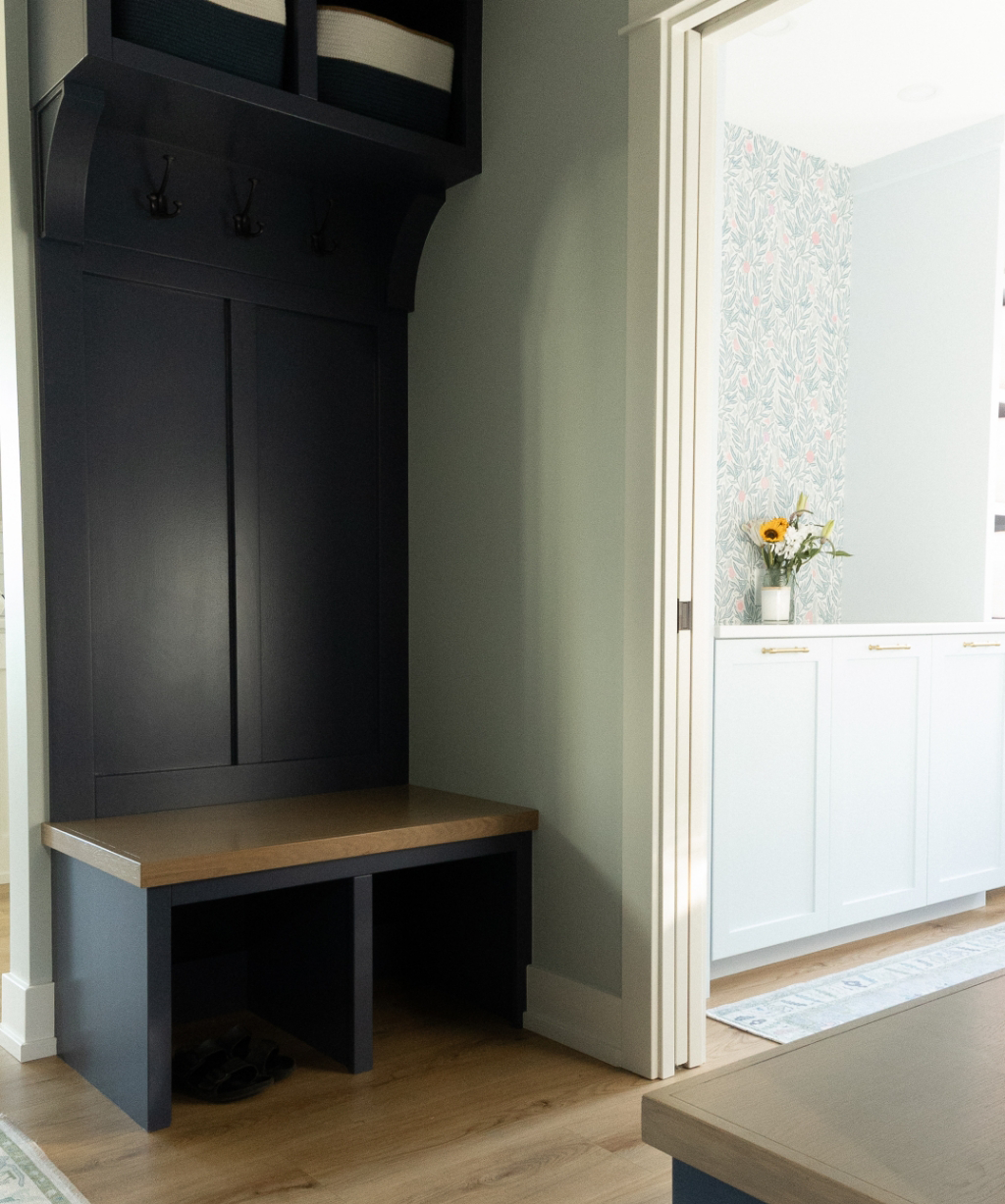 A built-in navy mudroom bench with hooks and storage, part of a Byron Center Whole Home Remodel, sits beside a doorway leading to a bright room with floral wallpaper and white cabinets.