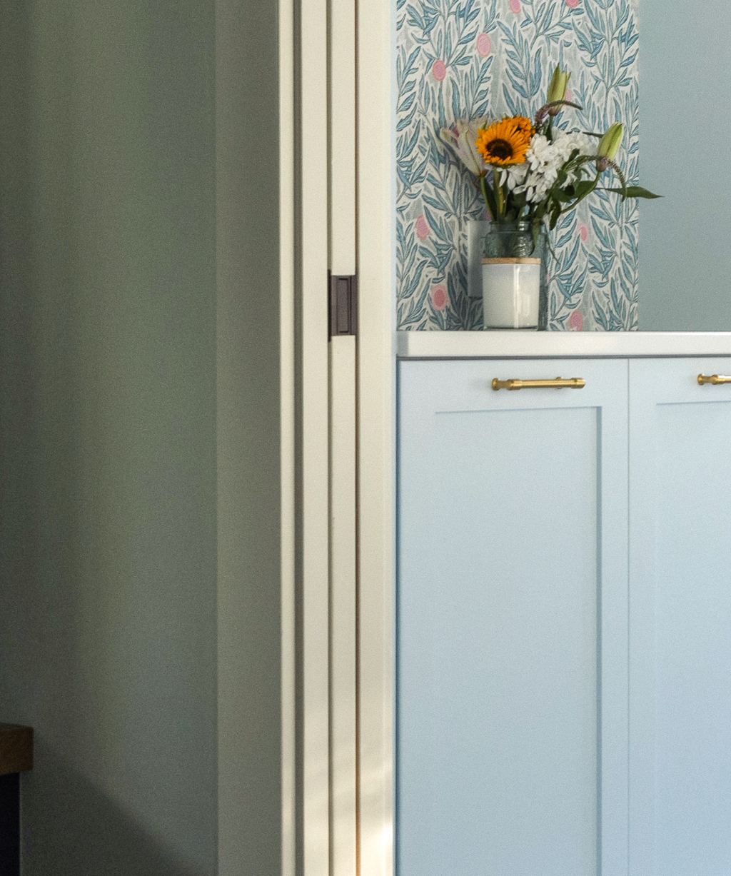 A jar of sunflowers and other flowers sits on a light blue cabinet with gold handles, part of a Byron Center Whole Home Remodel, against a floral patterned wallpaper.