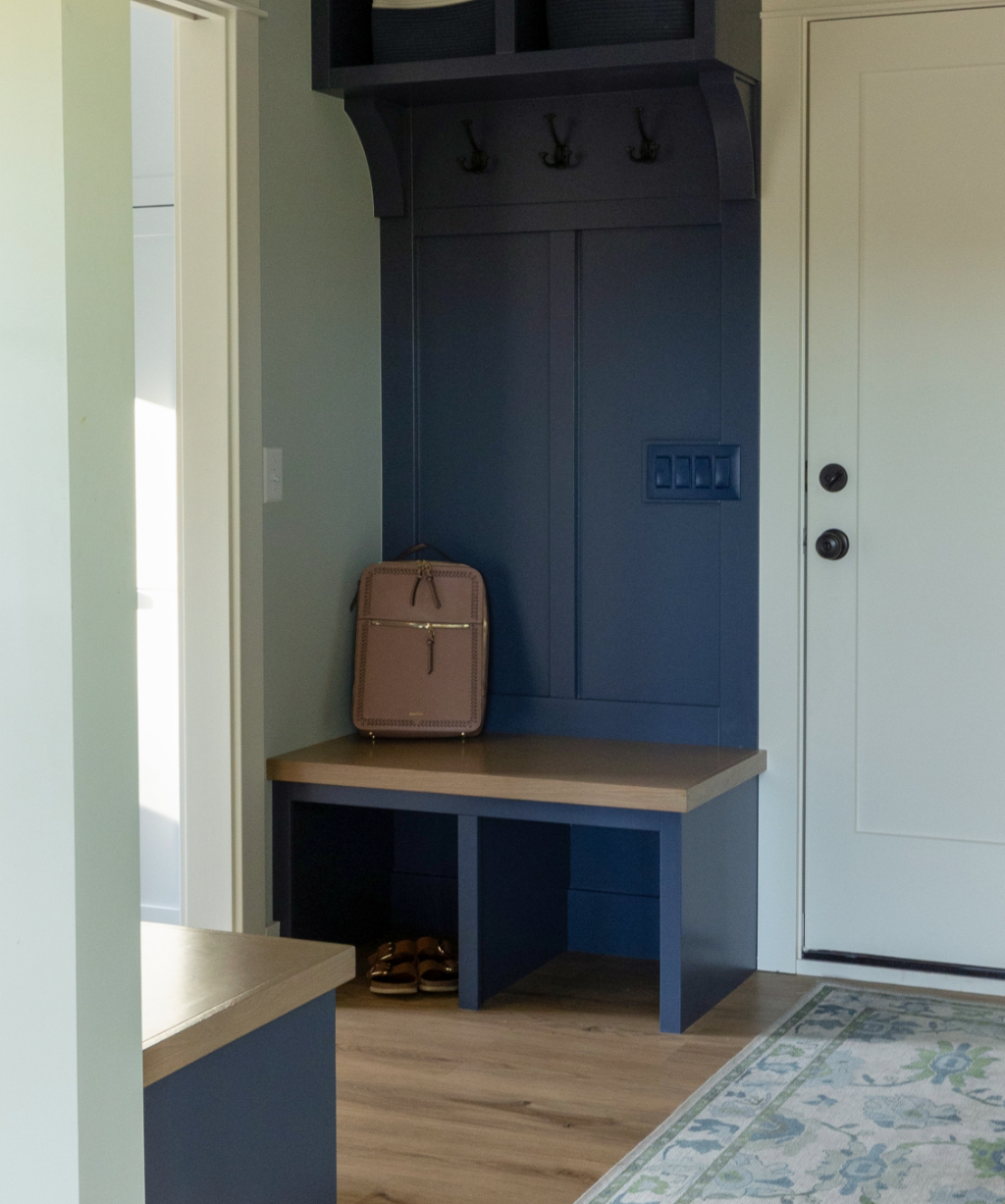 A brown backpack sits on a blue bench with cubby storage below and hooks above, next to a white door in a tidy entryway—part of a stunning Byron Center Whole Home Remodel.