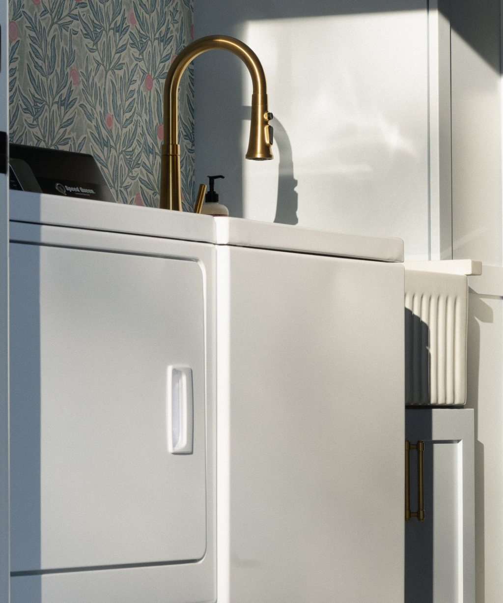 A gold faucet above a white sink with a soap dispenser, set against floral wallpaper and white cabinetry, brings elegance to this Byron Center Whole Home Remodel laundry room.