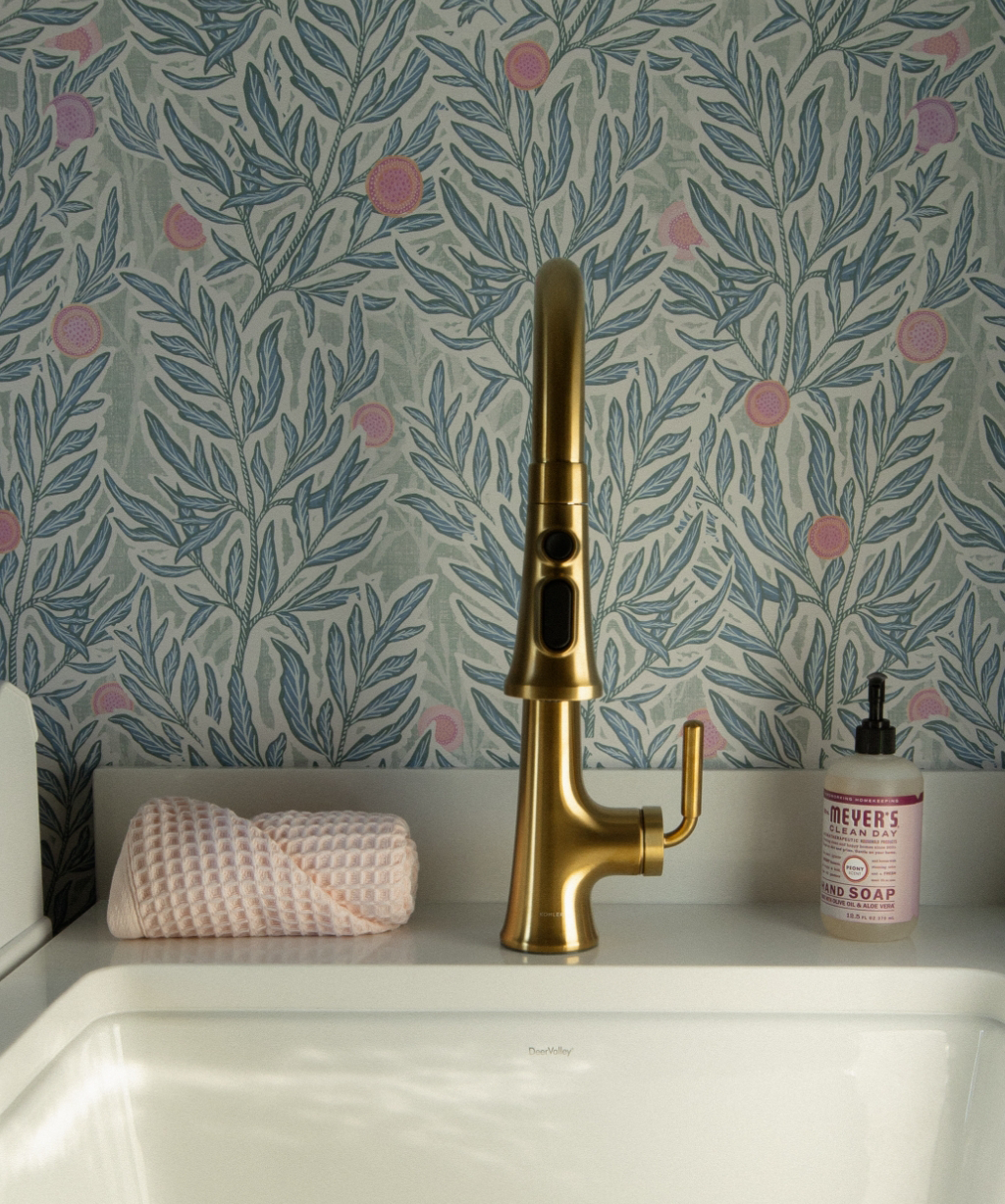 A gold faucet is mounted on a white sink with a pink textured towel and a bottle of Mrs. Meyer's hand soap, adding elegance to the Byron Center Whole Home Remodel, set against floral-patterned wallpaper.
