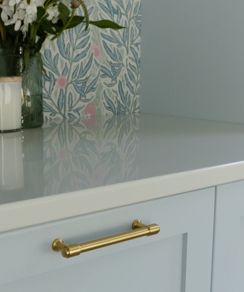 A white countertop with a gold drawer handle, a glass vase with flowers, a white candle, and blue floral wallpaper evoke the elegant style of a Byron Center Whole Home Remodel.