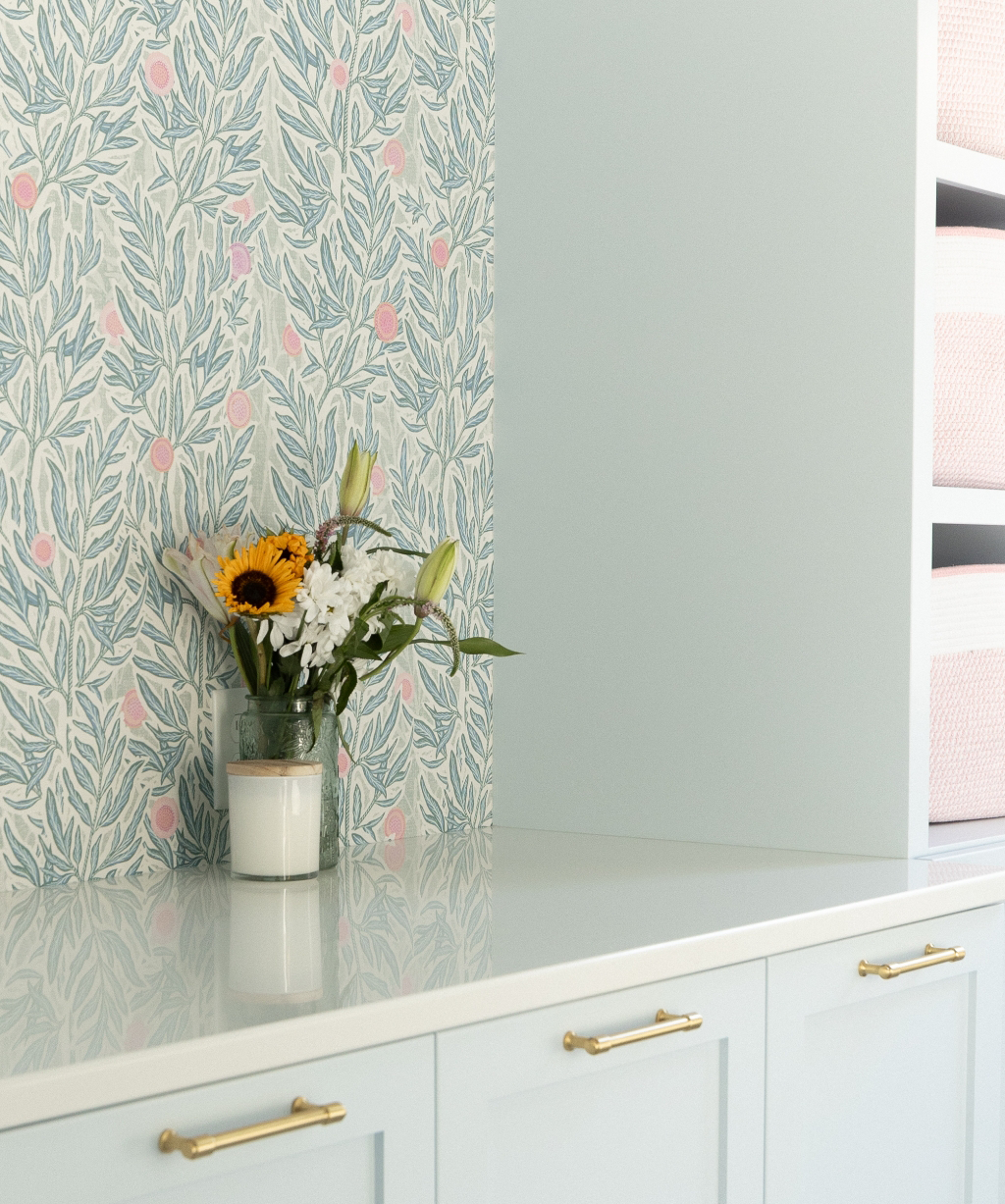 A vase with sunflowers and white flowers, and a candle sit on a white cabinet with gold handles—part of a stunning Byron Center Whole Home Remodel—next to a wall adorned with floral wallpaper.