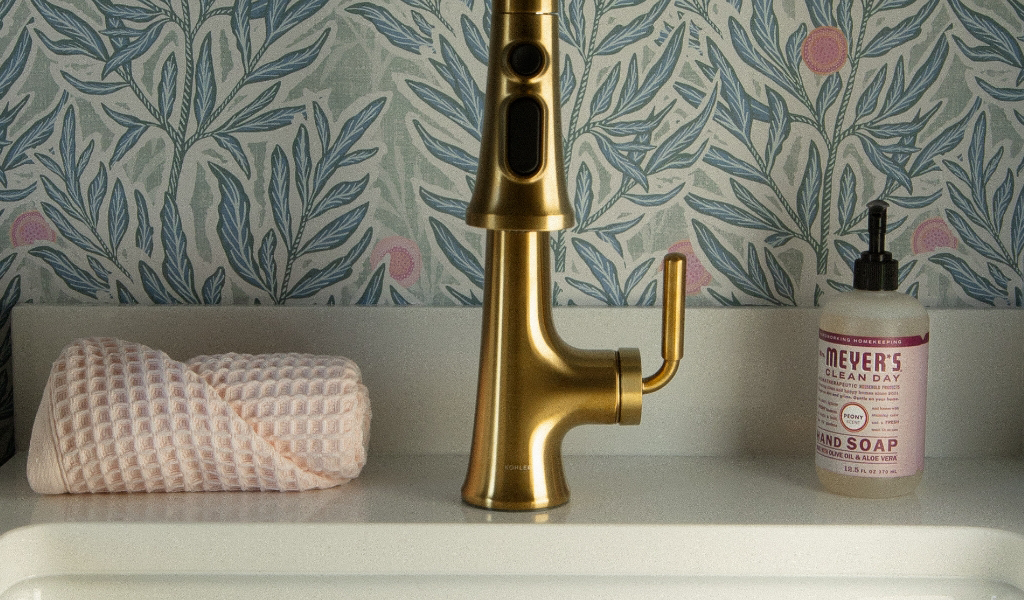 A gold faucet is centered above a white sink, with a pink textured towel on the left and a bottle of Mrs. Meyer's hand soap on the right, against a blue floral wallpaper background in a Byron Center remodel .
