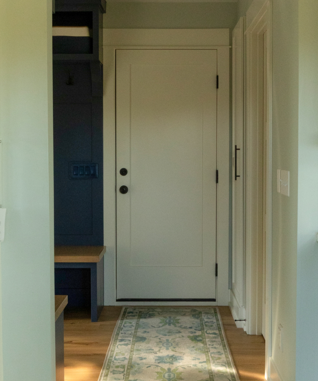 A closed white door at the end of a small hallway with light green walls, a patterned rug, and a built-in blue bench with storage showcases the elegance of this Byron Center Whole Home Remodel.