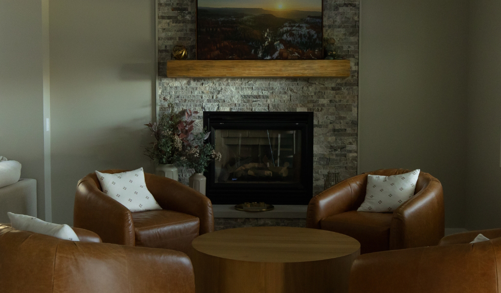 Four brown leather chairs with white pillows are arranged around a round wooden table in front of a stone fireplace, showcasing the inviting design of this Byron Center Whole Home Remodel. A landscape photo hangs above the mantel for a finishing touch.