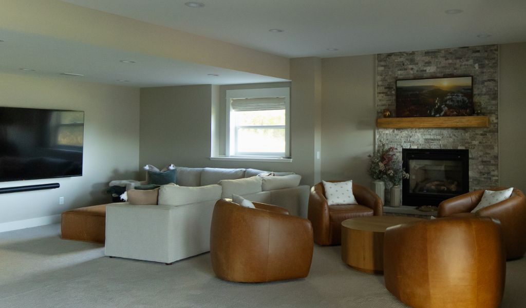 A modern living room from a Byron Center Whole Home Remodel featuring a sectional sofa, round leather chairs, a wall-mounted TV, and a fireplace with a stone surround and mantel.
