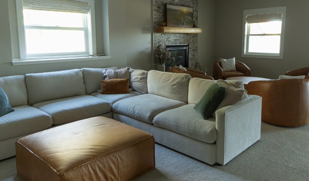 A modern living room from a Byron Center Whole Home Remodel, featuring a large beige sectional sofa, brown leather ottoman, matching armchairs, a stone fireplace, and elegant neutral decor.