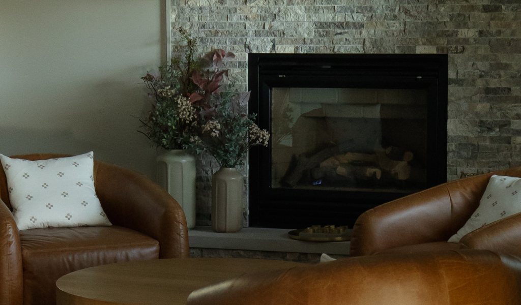 Two brown leather chairs with white patterned pillows face a round wooden table in front of a stone fireplace and vases with dried flowers, showcasing the inviting style of this Byron Center Whole Home Remodel.
