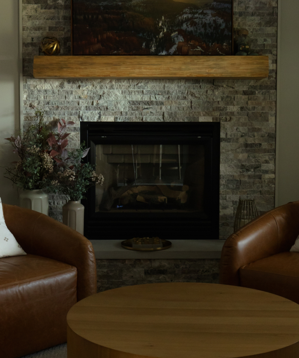 A modern living room from a Byron Center Whole Home Remodel featuring a stone fireplace, wooden mantel, dried floral arrangement, and two brown leather chairs around a round wooden coffee table.