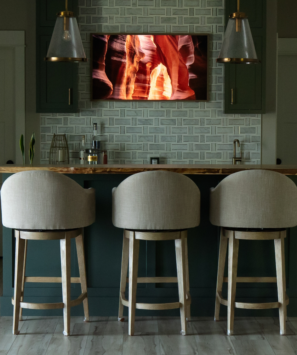 A modern kitchen from a Byron Center Whole Home Remodel, featuring three beige barstools, a green island, pendant lights, and a wall-mounted TV displaying a canyon scene.