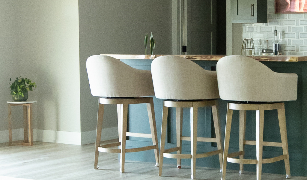 Three upholstered bar stools are lined up at a kitchen island with a light wood countertop—a stylish detail from this Byron Center Whole Home Remodel. A small plant sits on a side table in the background, adding a touch of greenery.