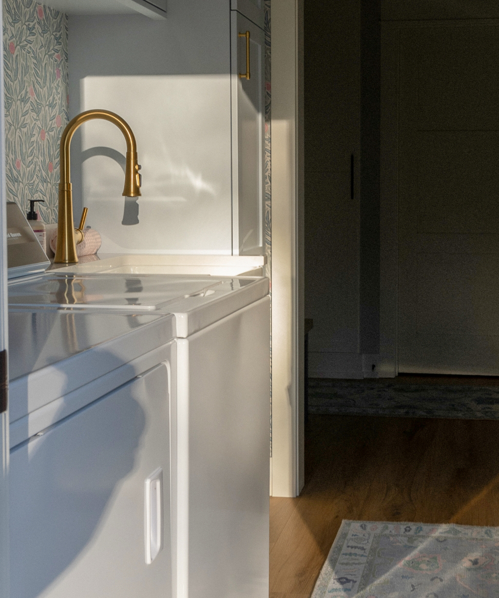 Sunlight falls on a white laundry sink with a gold faucet, reflecting on the cabinet. In this Byron Center Whole Home Remodel, wallpaper, part of a rug, and wooden floors complete the brightly lit room.