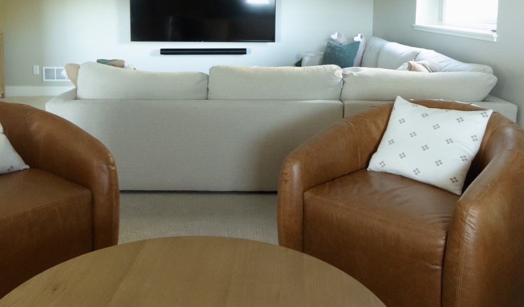 Satkoski – 40 A living room in this Byron Center Whole Home Remodel features two brown leather chairs, a round wooden coffee table, a beige sofa, and a wall-mounted TV.